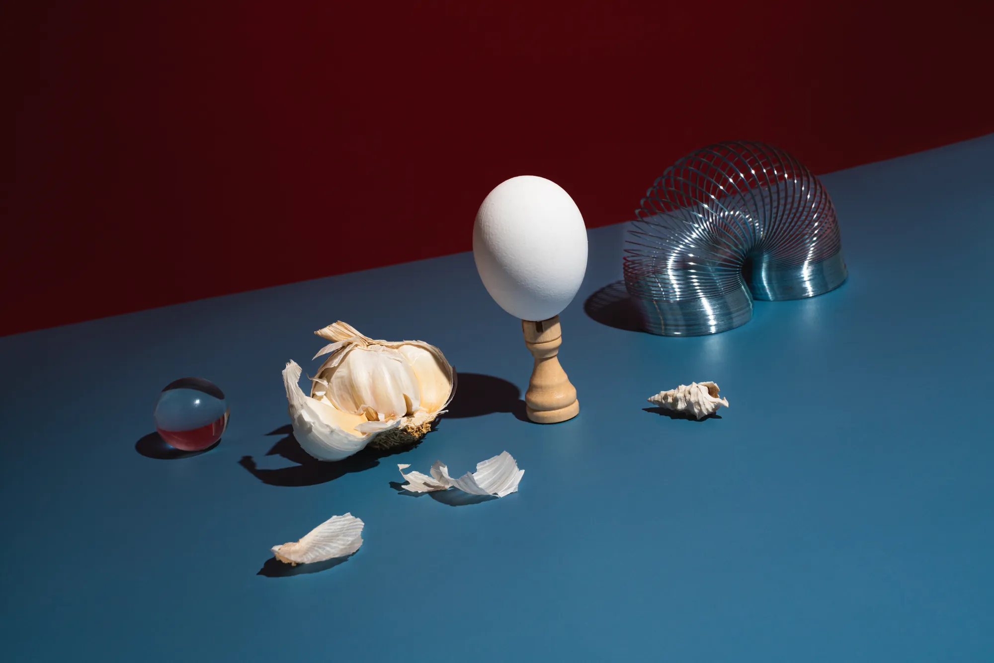 An egg on a wooden stand beside garlic, shells, a marble, and a Slinky on blue and red