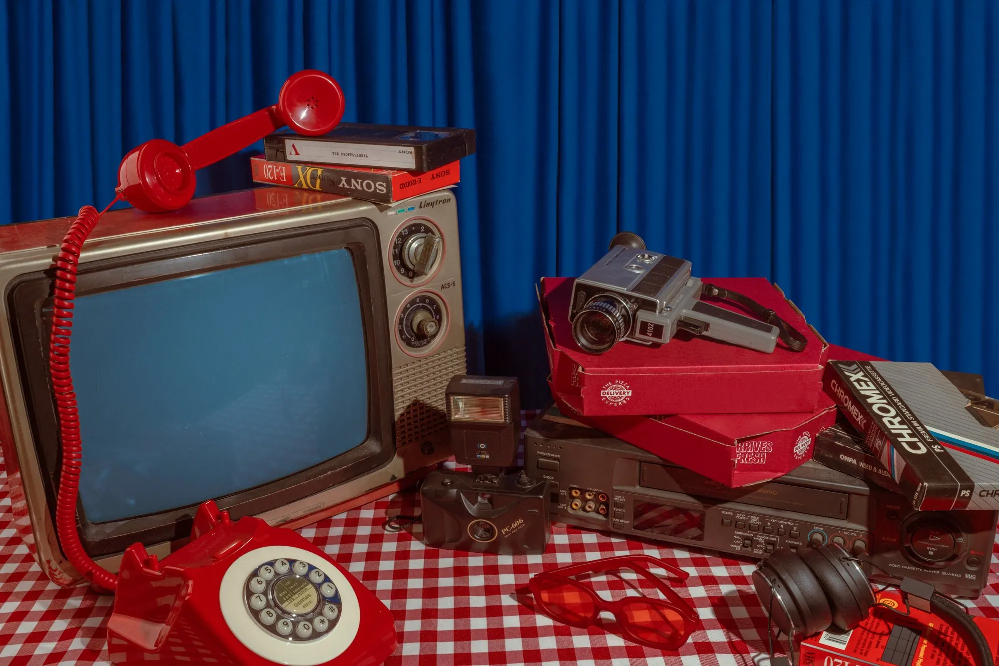 A vintage TV, red phone, and cameras on gingham with a blue curtain